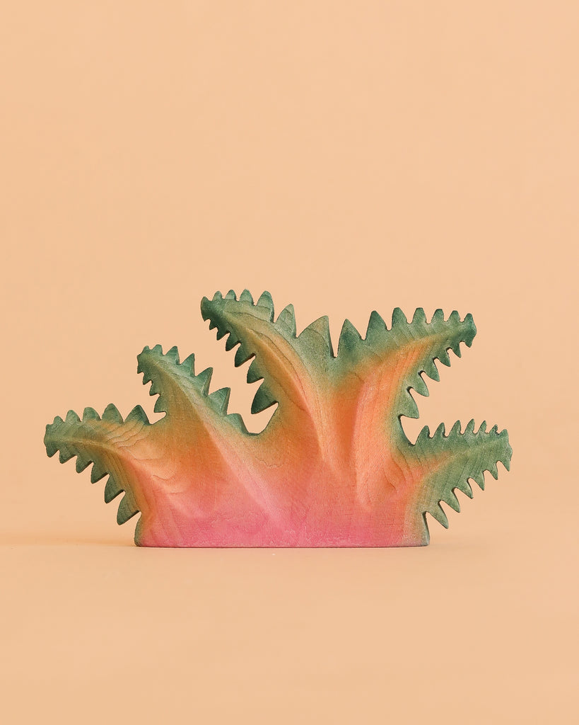 The Bumbu Handmade Wooden Fern Bush is a handcrafted wooden toy, painted in pink and green, with abstract spiky shapes reminiscent of fern foliage, pictured against a plain peach background.