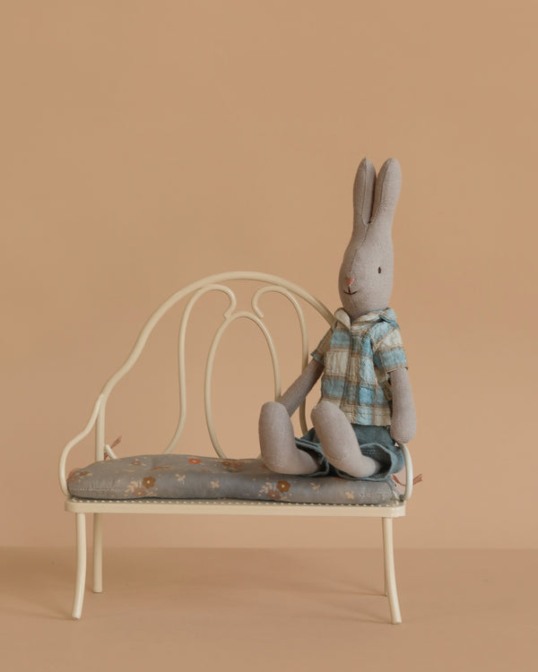 The Maileg Rabbit, Size 1 - Classic, wearing a plaid shirt and shorts, sits upright on a vintage-style metal bench with a patterned cushion against a plain beige background.