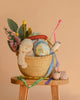 The Easter Basket Gift Set, filled with a plush bunny, decorative eggs, felt flowers, and colorful ribbons, sits on a wooden stool with two small fabric birds nearby against a warm beige background.