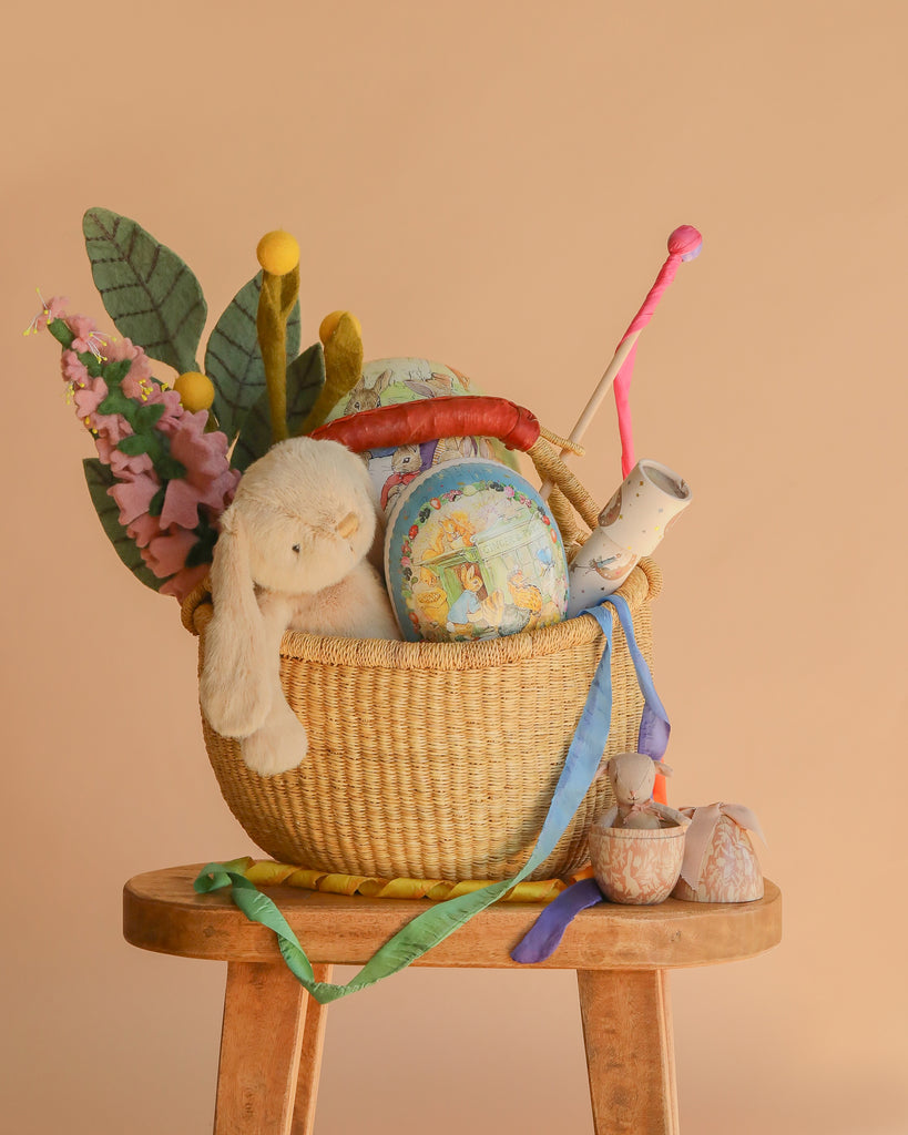 The Easter Basket Gift Set, filled with a plush bunny, decorative eggs, felt flowers, and colorful ribbons, sits on a wooden stool with two small fabric birds nearby against a warm beige background.