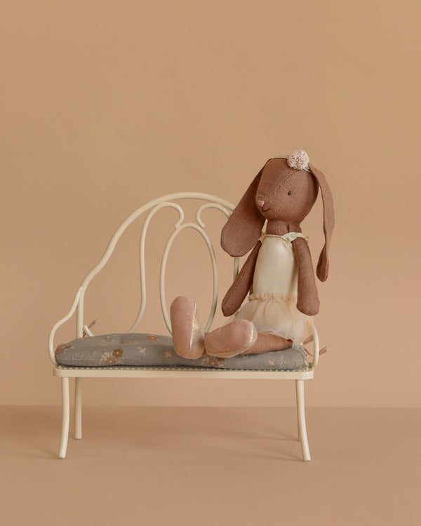 The Maileg Bunny, Size 2 in chocolate brown, dressed in a cream ballet suit and skirt, sits gracefully on a small white bench with a gray seat against a plain beige background.