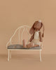The Maileg Bunny, Size 2 in chocolate brown, dressed in a cream ballet suit and skirt, sits gracefully on a small white bench with a gray seat against a plain beige background.