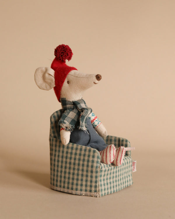 The Maileg Christmas Mouse - Big Brother, crafted from soft cotton, is adorned with a red hat and scarf as it relaxes in a green checkered armchair. It wears blue overalls and red-and-white striped socks, set against a soft beige background.