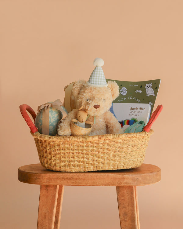 The Easter Basket Gift Set sits on a wooden stool, holding a teddy bear in a party hat, a small plush duck, wrapped gifts, colored pencils, and a Kids Drawing Book against a plain peach background.