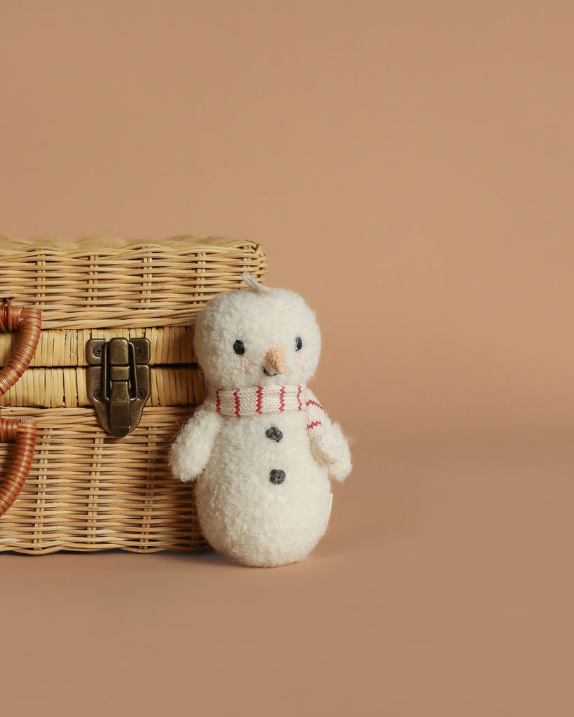 The Cuddle + Kind Baby Snowman - Candy Cane, featuring a red and white striped scarf, stands by a wicker basket on a beige background—an ideal collectible plush for stuffed animal fans.