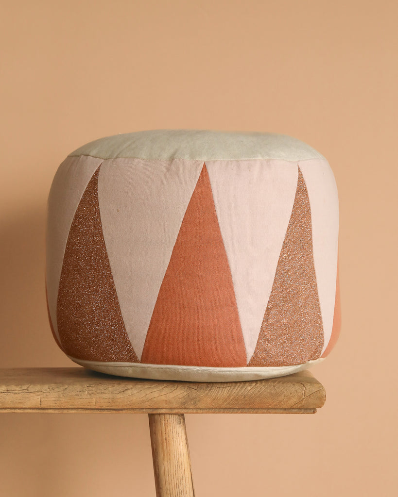 The Maileg Drum Pouf, featuring a round shape and a geometric pattern of salmon and rust-colored triangles, rests on a wooden bench and adds stylish accents to your space against a beige background.