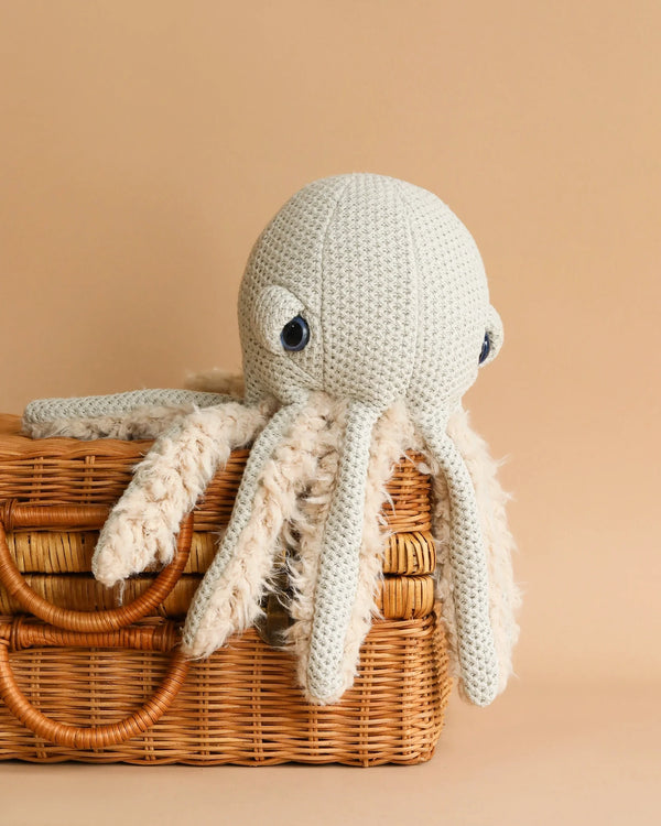 A BigStuffed Mini Grandma Octopus with light grey yarn and blue eyes rests atop a woven wicker basket against a beige background. The toy's tentacles dangle over the edge of the basket, offering a cozy, handmade feel. Perfect as a children's toy, this mini plush octopus adds charm to any nursery.
