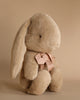 The Maileg Medium Plush Bunny in Dusty Brown sits upright against a neutral beige background, featuring long ears and polyester fillings. It boasts a soft texture and is adorned with a pink bow around its neck.