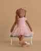 A Maileg Bunny, Size 3 in chocolate brown, dressed in a rose ballet suit and skirt, sits cross-legged on a small white bench against a beige background.