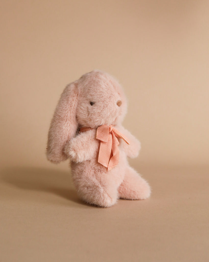 The Maileg Mini Plush Bunny - Powder, crafted from a soft fabric and filled with polyester, features long ears and a matching pink ribbon around its neck as it sits against a warm beige background.