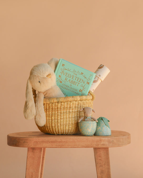 The Easter Basket Gift Set, featuring a plush rabbit, a Maileg Easter Egg, and "The Velveteen Rabbit" book, sits on a wooden stool with two decorative objects beside it against a beige background, evoking classic childhood tales.