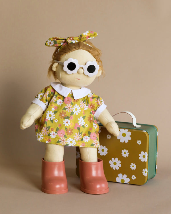 A Dinkum Doll - Poppet (Extended Pack) with button eyes and blonde yarn hair, wearing a removable unisex outfit, standing beside a small green suitcase against a beige background.
