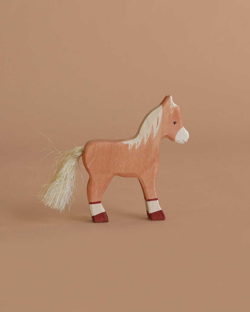 A handcrafted Holztiger Foal, Standing features a light brown body, white mane and tail, and red leg accents. Made in Europe with painted details, this wooden toy stands on a neutral beige background.