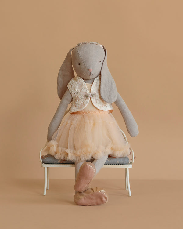 The Maileg Bunny, Size 3 - Classic - Ballet Suit and Skirt - Cream, sits on a small white bench in a peach tulle skirt, sequin vest, and ballet slippers against a plain beige background.