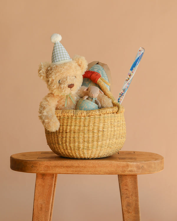 The Easter Basket Gift Set, featuring a teddy bear in a party hat, a plush mouse, Snuggle Bunnies, a toy mushroom, and a Glitter Magic Wand, sits on a wooden stool against a plain beige background.