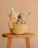 The Easter Basket Gift Set, featuring a teddy bear in a party hat, a plush mouse, Snuggle Bunnies, a toy mushroom, and a Glitter Magic Wand, sits on a wooden stool against a plain beige background.