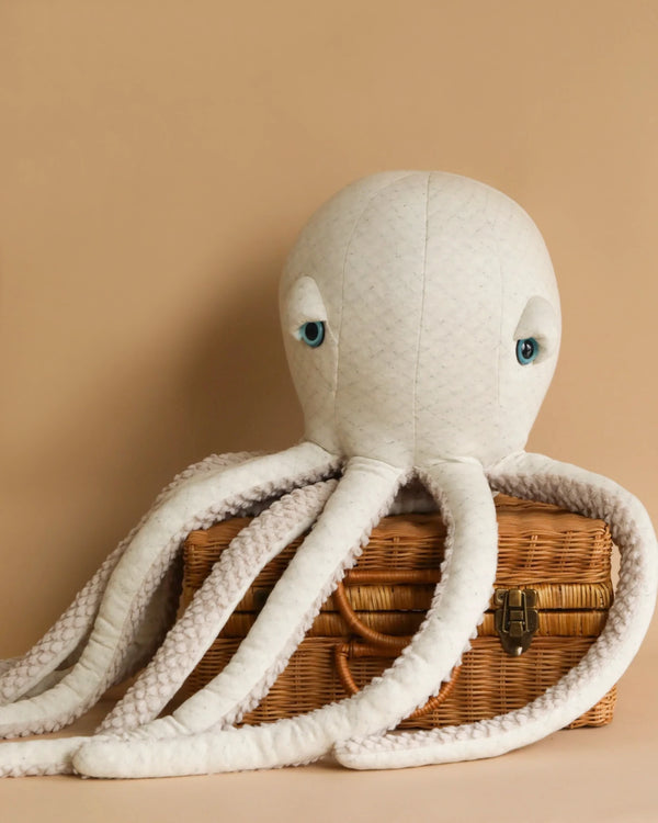 A BigStuffed Octopus - Big Albino with white, textured fabric and blue eyes is draped over a small wooden basket with a latch, set against a beige background. The octopus's tentacles hang down around the basket, evoking an albino octopus exploring its underwater world.