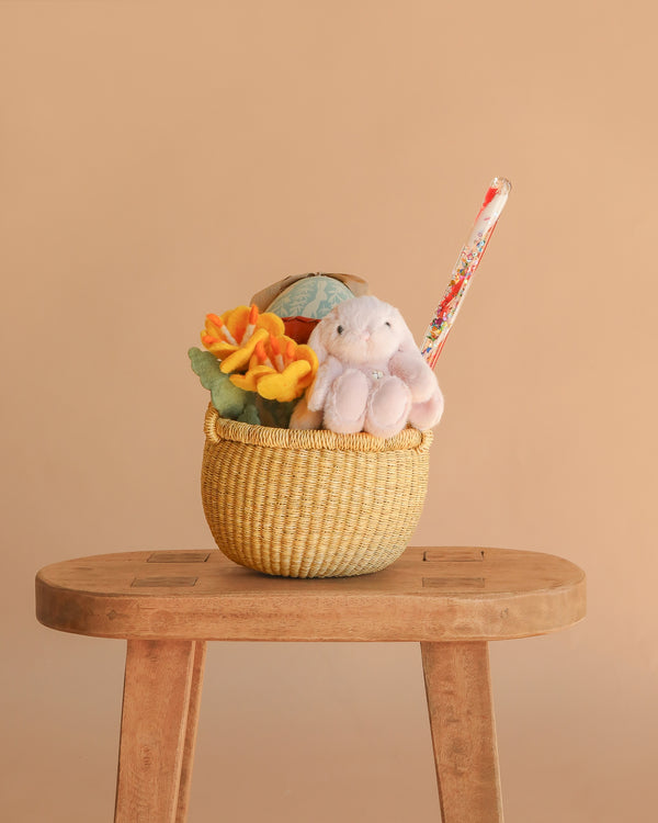 An Easter Basket Gift Set, featuring a plush bunny, felt flowers, and a decorative tube, sits on a wooden stool against a plain beige background.