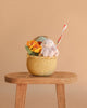 An Easter Basket Gift Set, featuring a plush bunny, felt flowers, and a decorative tube, sits on a wooden stool against a plain beige background.
