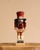 The Christian Ulbricht Collectible Wooden Nutcracker - Toy Trader features a tall brown hat, bushy beard, red coat, and beige shorts. It stands on a wooden surface against a beige background, holding a small decorated box.