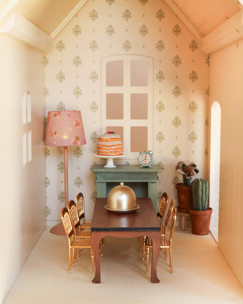 Experience a beautifully arranged miniature dining room featuring a table, six gold chairs, a covered dish, a cake, a lamp, a clock, plants, and patterned wallpaper—all part of the elegant Maileg Fully Furnished Dollhouse Set + Gift With Purchase (restock expected in August), complete with charming dollhouse furniture for your perfect Maileg home.