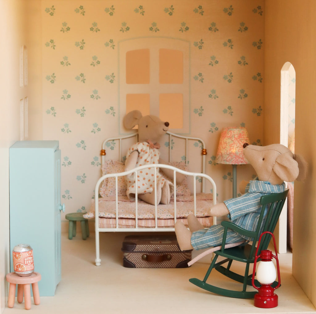 Discover the Maileg Fully Furnished Dollhouse Set + Gift With Purchase (Restock Expected In August), featuring two fabric mice in a miniature bedroom—one nestled on a bed with polka-dot bedding, the other relaxing in a rocking chair. The room is styled with charming dollhouse furniture, including a lamp, suitcase, cabinet, pink stool, can, and lantern. Delicate wallpaper decorated with tiny blue flowers adds to the cozy ambiance of this enchanting Maileg dollhouse scene.