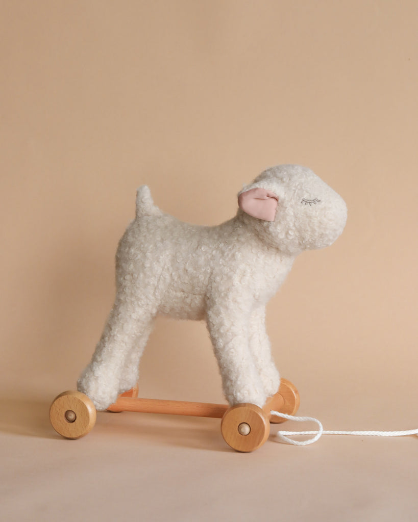 A plush toy sheep with fluffy white wool, mounted on a wooden wheeled base with a pull string, stands against a soft beige background, designed to enhance motor skills as a Pull-Along Mary Lamb.