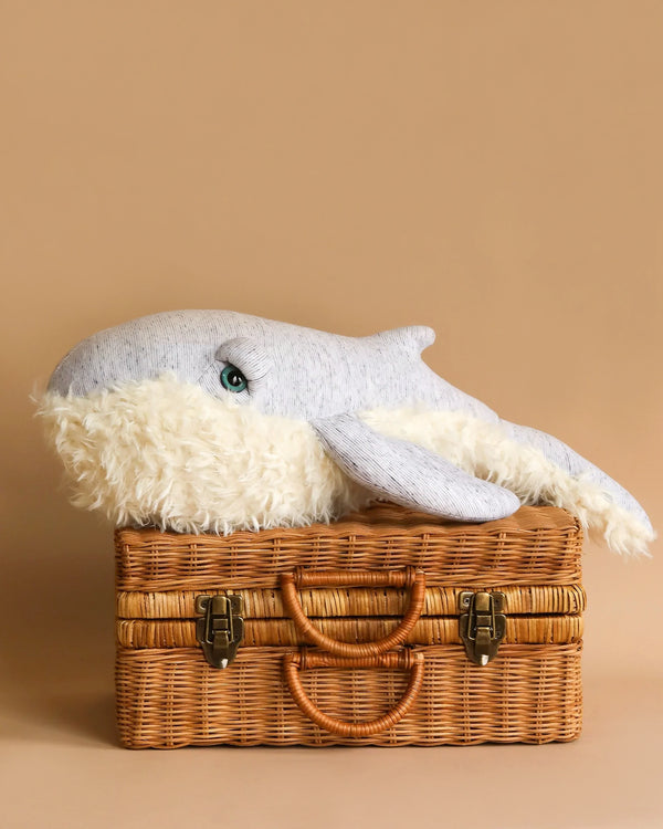 A BigStuffed Small Grandpa Whale with a blue and white body rests on top of a closed, light brown wicker suitcase with two clasp closures and two woven handles. This adorable cuddle companion is set against a solid beige background.