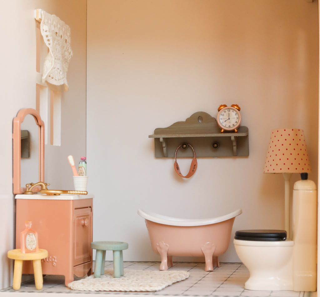 Discover the Maileg Fully Furnished Dollhouse Set + Gift With Purchase (Restock Expected In August), featuring a charming miniature bathroom scene with dollhouse furniture, including a pink tub, black and white toilet, sink, small stools, and a shelf with a clock. This adorable space is completed by a wall lamp, towel, and soft white rug—perfect for any Maileg dollhouse or other miniature displays.