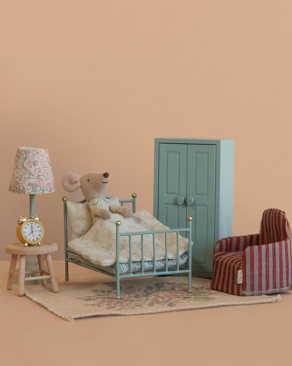 The Maileg Little Brother Mouse Bedroom Set features a mouse resting in a vintage bed with floral bedding, surrounded by Maileg furniture—a striped red armchair, teal wardrobe, lamp-topped stool, and clock—on a plain beige background.