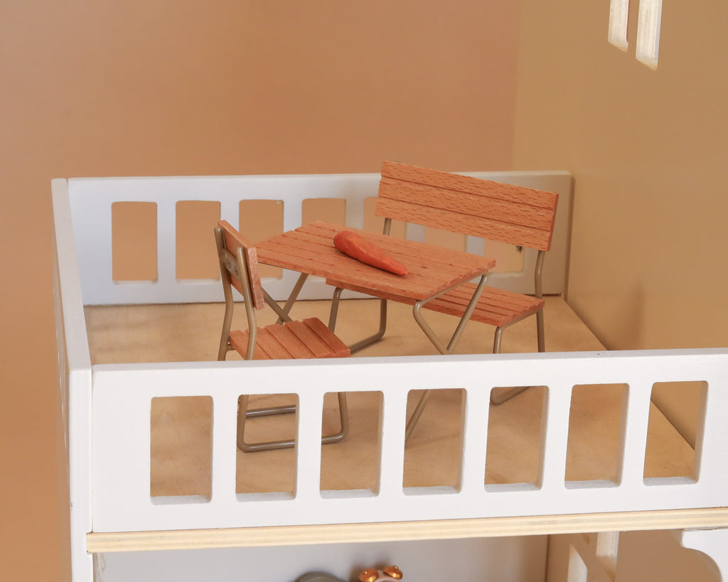 Bring your Maileg dollhouse to life with the Maileg Fully Furnished Dollhouse Set + Gift With Purchase (Restock Expected In August), featuring a delightful miniature patio scene complete with elegant dollhouse furniture—including a small wooden table, two chairs, a bench, and a single carrot placed atop the table inside a charming white balcony model—perfect for enhancing any miniature dollhouse setting.