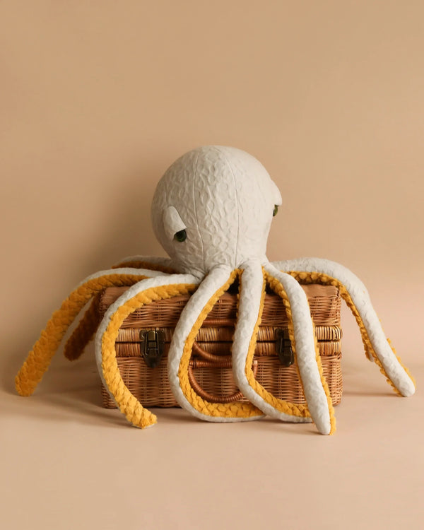 A BigStuffed Octopus - Pop with a white head and long, yellow tentacles is draped over a closed, brown wicker basket. Made in Lithuania, it evokes the charm of the underwater world against a solid light beige background.