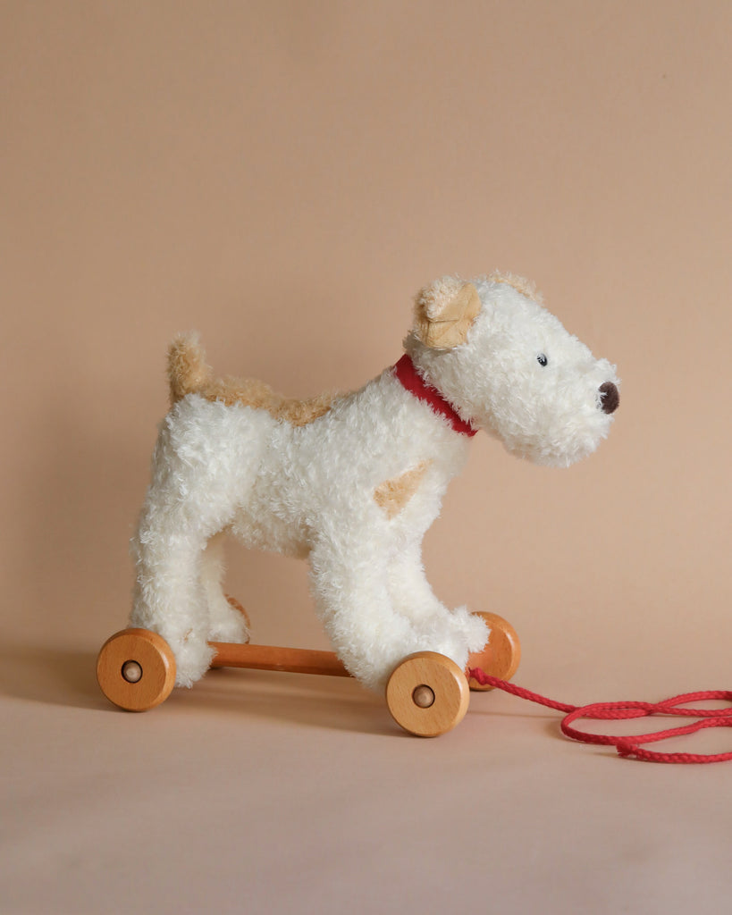 A Pull-Along Eliot Dog featuring a stuffed dog with white fur and a red collar on wooden wheels, connected by a red string, designed against a light peach background to enhance motor skills.