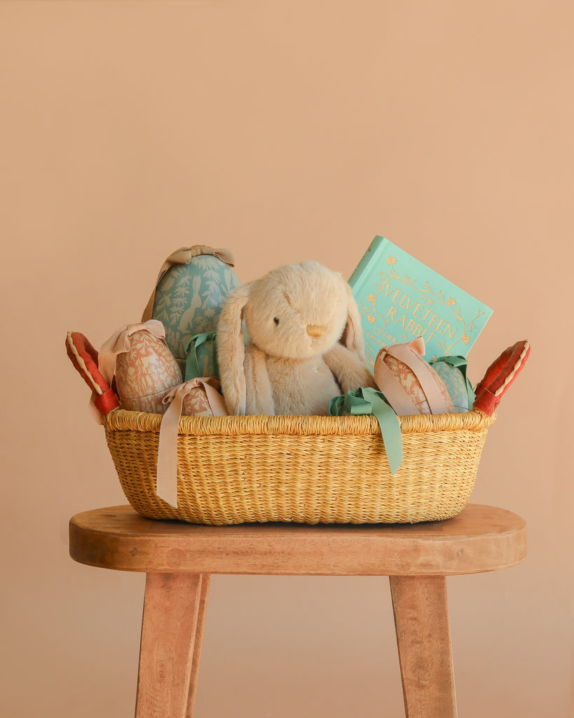 An Easter Basket Gift Set with a plush bunny, soft toys, and a children’s book rests on a wooden stool against a beige background—a charming seasonal display.