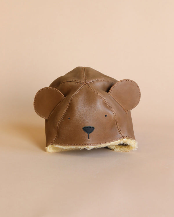 A brown Donsje Leather Classic Hat - Bear shaped like a bear's head, with round ears and a sheep wool lining, set against a soft beige background.