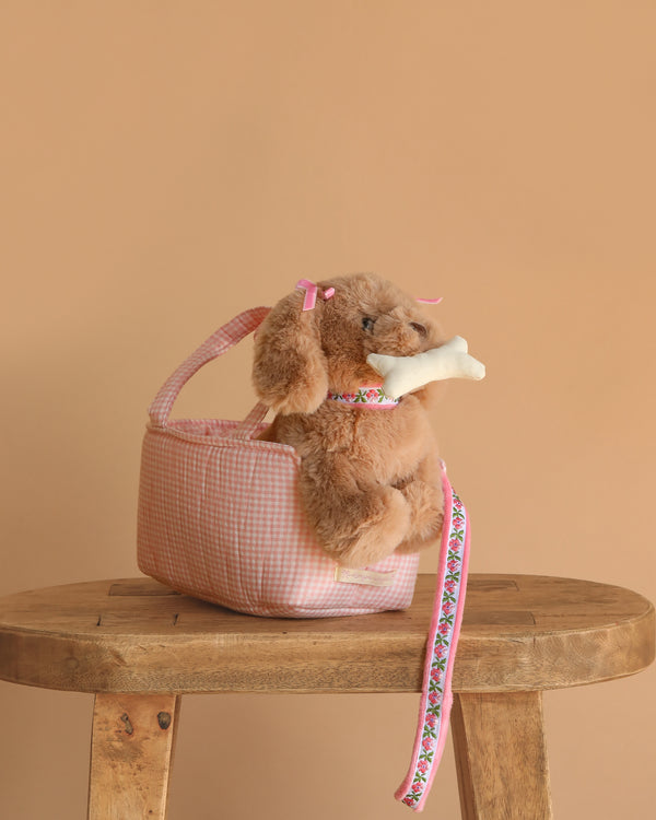 The Konges Sloejd Maude Puppy in Basket features a soft toy puppy with pink bows and a bone, nestled in a pink gingham basket with a floral leash, displayed on a wooden stool against a beige background.