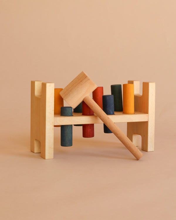 A Rainbow Pound -a- Peg wooden Montessori toy bench with red, yellow, blue, green, and orange pegs and a wooden mallet resting on top, set against a plain beige background. This eco-friendly toy encourages colorful play and learning.