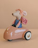 A Maileg Sister Mouse Holiday Adventure Set in a denim dress and headscarf sits in a small pink car with a rolled blanket tied to the back, set against a soft beige background.