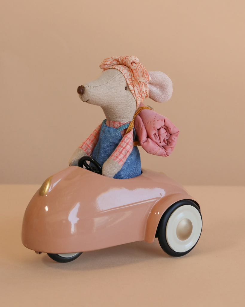 A Maileg Sister Mouse Holiday Adventure Set in a denim dress and headscarf sits in a small pink car with a rolled blanket tied to the back, set against a soft beige background.