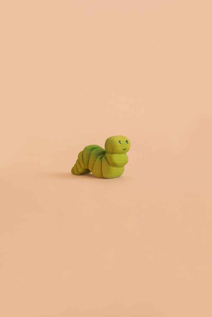 A small, green Bumbu Handmade Wooden Caterpillar with a cute, smiling face sits on a plain peach background, evoking the charm of a Montessori toy perfect for imaginative play.