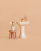 A Maileg Powder Room Set features a stool holding a cup with two toothbrushes and a toothpaste tube beside a bathroom sink with a gold faucet and two bottles, all arranged against a beige background.