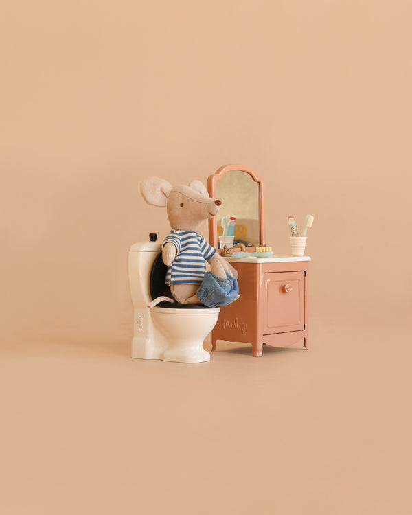 A Maileg LouiMax Bathroom Set features a mouse in a striped shirt and shorts sitting on a toilet beside a charming sink dresser with mirror, toothbrushes, and toothpaste, all set against a plain beige background.