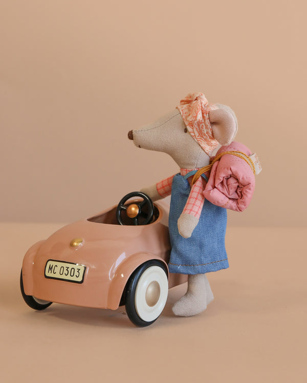 The Maileg Sister Mouse Holiday Adventure Set features a mouse in a headscarf, plaid shirt, and denim dress with a sleeping bag, standing beside a pink Maileg Mouse Car against a plain beige background.
