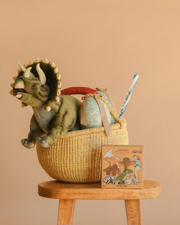 The Easter Basket Gift Set, featuring a Triceratops Dinosaur Stuffed Animal, a blue-patterned item, and a vintage-style illustrated box, sits on a wooden stool against a beige background.