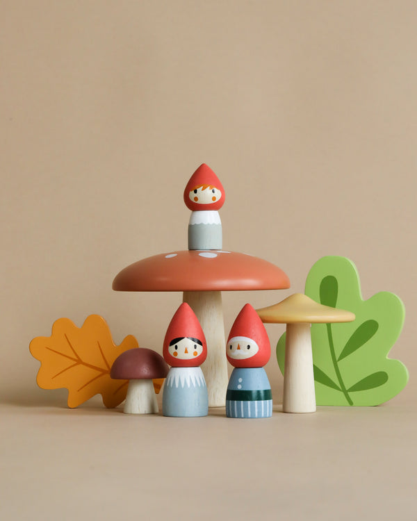 A whimsical Woodland Gnome Family scene from the Merrywood Tales collection featuring mushroom-shaped houses and figures with three leaf accessories on a beige background.