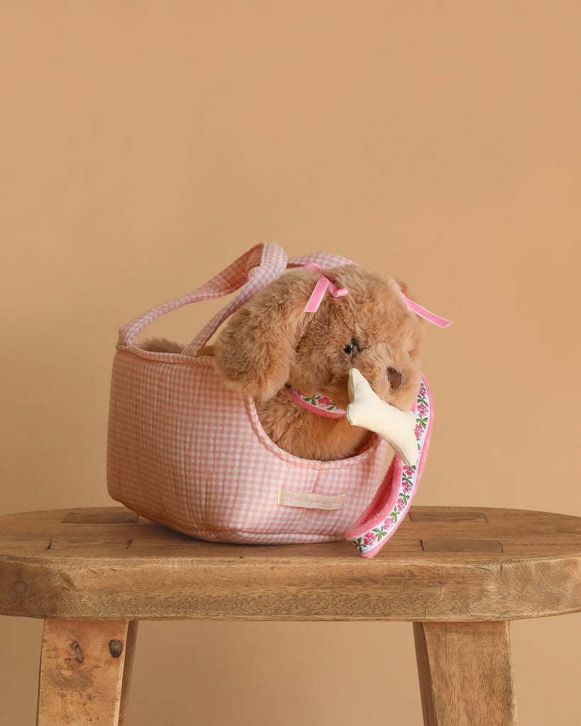 The Konges Sloejd Maude Puppy in Basket sits with pink bows and a floral toy bone in its mouth, nestled in a pink gingham carrier on a wooden bench against a beige background.