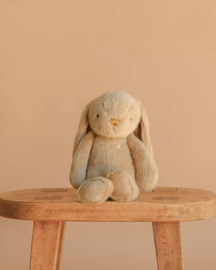 The Large Easter Basket Gift Set features a soft, beige stuffed bunny from the Snuggle Bunnies collection sitting upright on a wooden stool against a plain light brown background.
