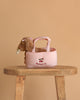 The Konges Sloejd Maude Puppy in Basket features a plush puppy with a pink bow inside a pink gingham dog basket embroidered with cherries and "cherries out," displayed on a wooden stool against a beige background.