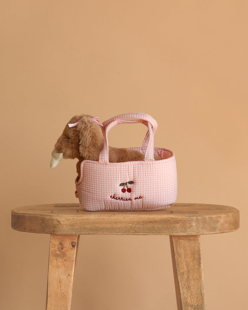 The Konges Sloejd Maude Puppy in Basket features a plush puppy with a pink bow inside a pink gingham dog basket embroidered with cherries and "cherries out," displayed on a wooden stool against a beige background.