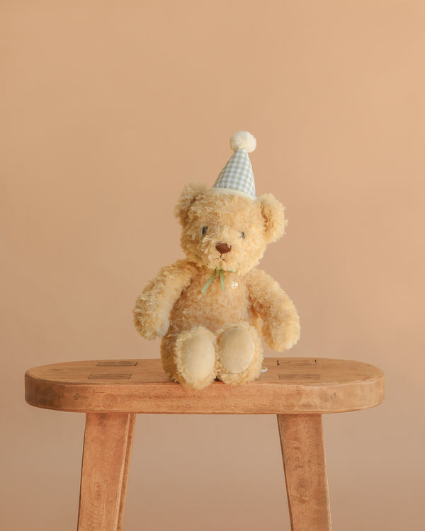 The Snuggle Bunnies - Birthday Arthur - Gingham is a plush toy bear in a blue and white party hat, seated on a wooden stool against a beige background—an ideal keepsake gift.